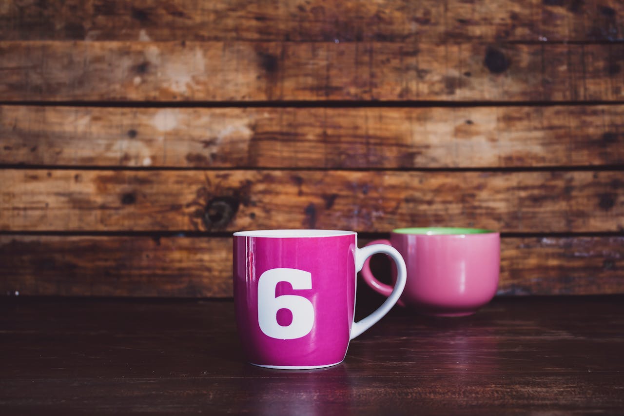 Bright pink ceramic mugs with number six design on a rustic wooden surface.