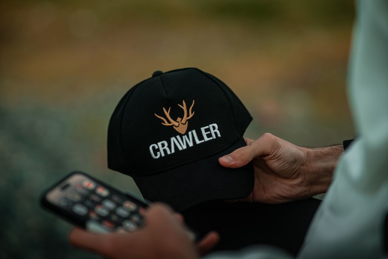 Close-up of a man's hand holding a cap with antler design and 'Crawler' text outdoors.