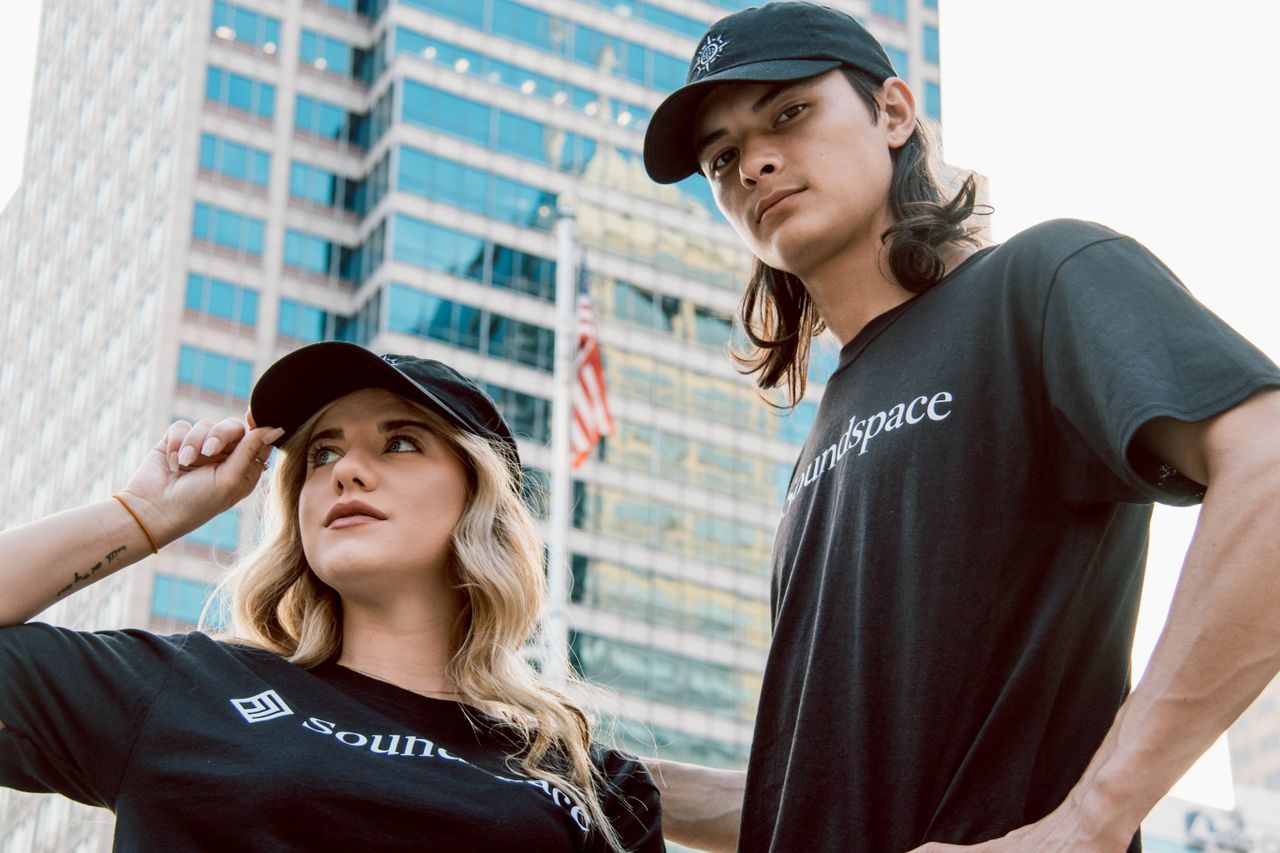 A fashionable couple wearing black t-shirts and caps in an urban setting with skyscrapers.